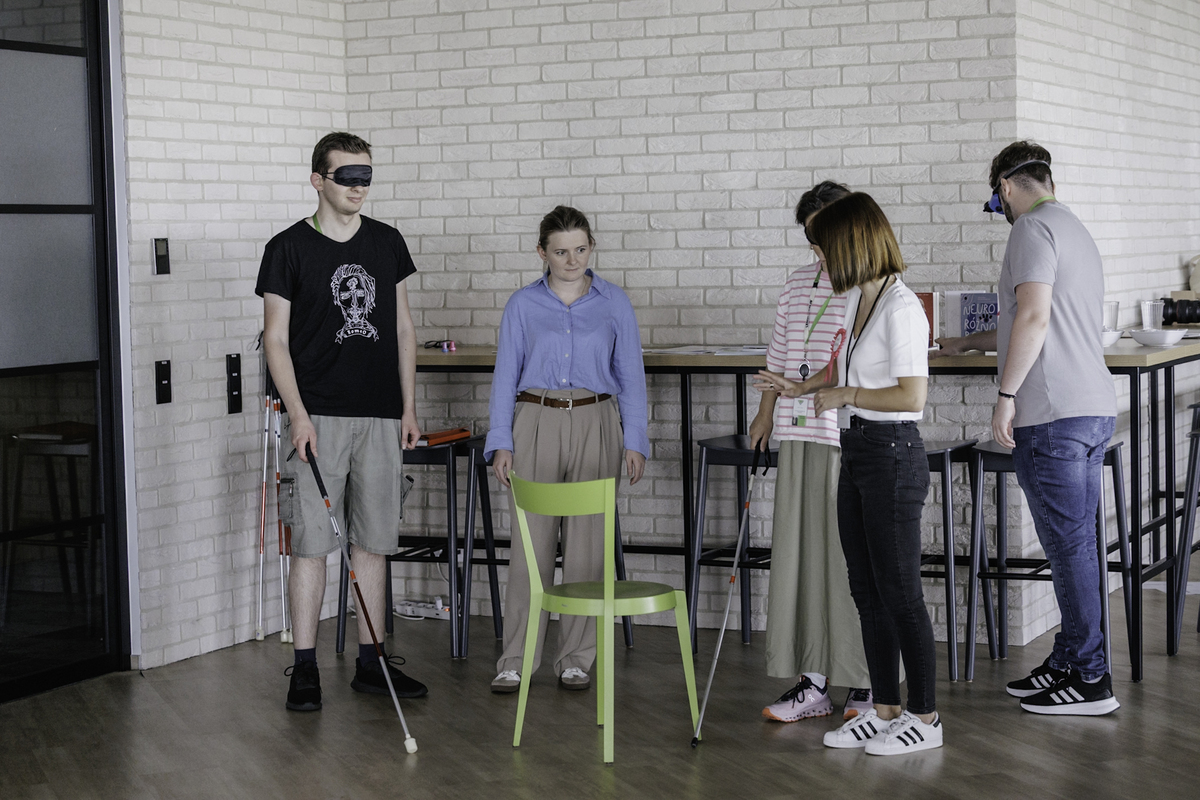 A group of five people participate in an exercise with white canes. Two people are blindfolded, the third stands in the middle. People from the Foundation team support the participants.